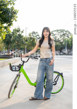 Pretty asian woman holding green rental bicycle standing on pavement in park or plaza under sunlight Pretty asian woman holding green rental bicycle standing on pavement in park or plaza under sunlight 136655535