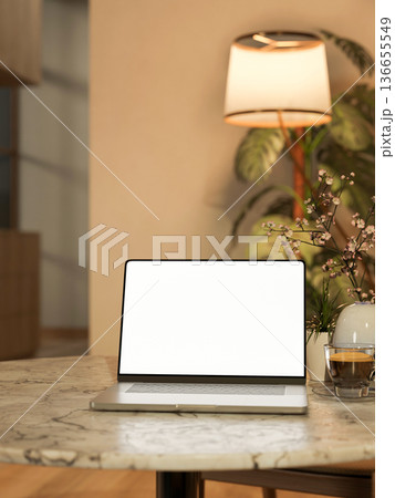 White screen laptop with coffee cup aside vase on round marble table across lamp in dim living room. White screen laptop with coffee cup aside vase on round marble table across lamp in dim living room. 136655549