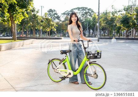 Laughing asian woman holding green rental bicycle standing on pavement in park plaza under sunlight. Laughing asian woman holding green rental bicycle standing on pavement in park plaza under sunlight. 136655881