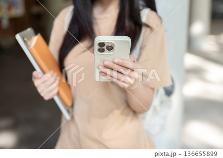 Close up of woman college student holding book looking at phone while standing walking in a building Close up of woman college student holding book looking at phone while standing walking in a building 136655899