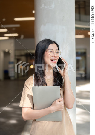 Glasses woman college student holding laptop talking on phone while standing or walking in building. 136655900