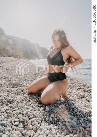Woman bikini beach kneeling on a pebbled shore with sunlight and ocean waves during summer vacation 136656080