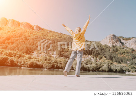 Woman, River, Mountains - Happy Woman with Arms Raised Standing on Riverbank with Mountains in Background. Woman, River, Mountains - Happy Woman with Arms Raised Standing on Riverbank with Mountains in Background. 136656362