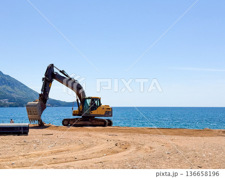 A yellow excavator standing on sandy beach near the sea and mountains. Industrial presence, construction and human activity by the coastline. A yellow excavator standing on sandy beach near the sea and mountains. Industrial presence, construction and human activity by the coastline. 136658196