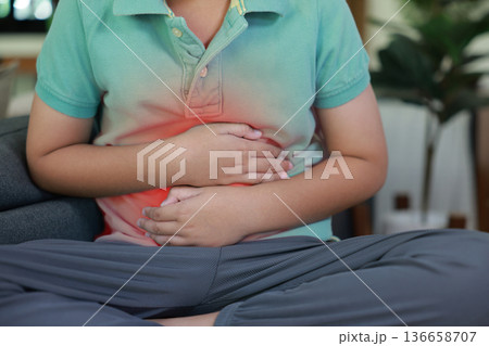 Asian boy holding belly suffering from severe abdominal pain with red inflamed area. Concept of stomach ache, indigestion, food poisoning, gastritis, and pediatric medical healthcare treatment. Asian boy holding belly suffering from severe abdominal pain with red inflamed area. Concept of stomach ache, indigestion, food poisoning, gastritis, and pediatric medical healthcare treatment. 136658707
