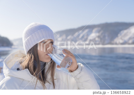 A woman stands by a river in winter and drinks from a glass filled with clear water. Snow covers the ground, and mountains are visible in the background. High quality photo A woman stands by a river in winter and drinks from a glass filled with clear water. Snow covers the ground, and mountains are visible in the background. High quality photo 136658922