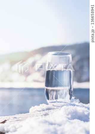 Water filled glass rests on fresh snow near a natural spring. Winter landscape surrounds the glass. Light reflects off the water. Nature looks bright and inviting in the cold weather. High quality Water filled glass rests on fresh snow near a natural spring. Winter landscape surrounds the glass. Light reflects off the water. Nature looks bright and inviting in the cold weather. High quality 136658941