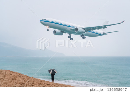 Flight trip to Phuket in Thailand during the rainy season. A plane flying low over the sea lands at the airport on the island, a vacationing tourist with an umbrella on the beach meets and photographs 136658947
