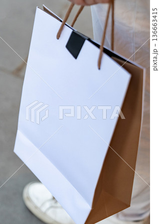 Person holding a blank paper shopping bag while walking outdoors. The clean surface allows flexible use for retail and commerce design. 136663415