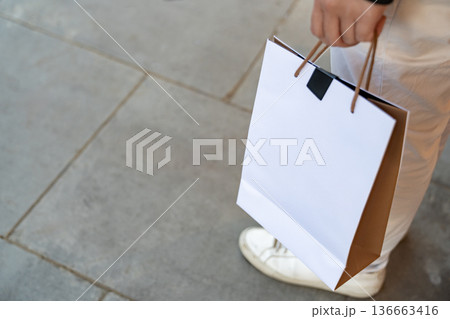 Person holding a blank paper shopping bag while walking outdoors. The clean surface allows flexible use for retail and commerce design. 136663416
