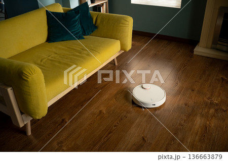 White robot vacuum cleaner autonomously navigating and cleaning a modern living room's parquet floor, offering smart home technology and automated household convenience White robot vacuum cleaner autonomously navigating and cleaning a modern living room's parquet floor, offering smart home technology and automated household convenience 136663879