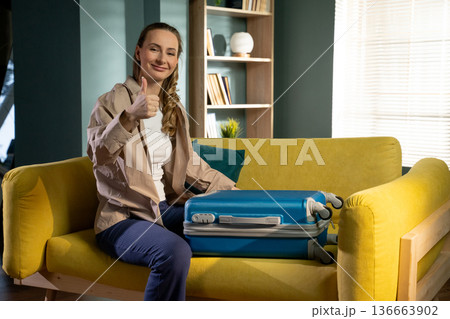 Woman sitting on a yellow sofa feeling excited and ready for a trip, giving a confident thumbs up gesture next to her packed blue suitcase, preparing for departure Woman sitting on a yellow sofa feeling excited and ready for a trip, giving a confident thumbs up gesture next to her packed blue suitcase, preparing for departure 136663902