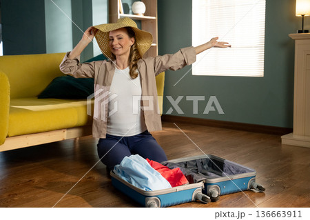 Happy woman wearing a straw hat and smiling broadly, sitting on the floor in her living room, packing clothes into an open suitcase, anticipating an exciting journey 136663911