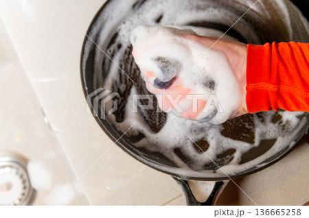 Hand in orange sleeve scrubbing a soapy black frying pan on a light countertop with bubbles and a sink drain visible in the background 136665258