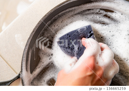 Hand scrubbing a cast iron skillet with a soapy sponge in a kitchen sink, bubbles and foam visible around the cookware on a light countertop Hand scrubbing a cast iron skillet with a soapy sponge in a kitchen sink, bubbles and foam visible around the cookware on a light countertop 136665259