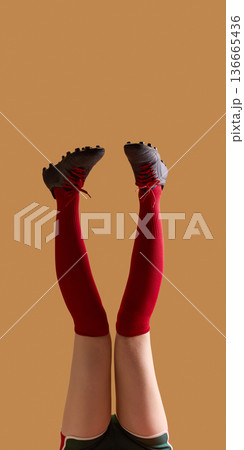 Female legs in red knee socks and black cleats vertical pose on beige background. 136665436