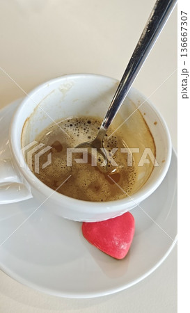 Cup of coffee with foam and spoon on saucer with heart shaped candy on table. Morning drink scene symbolizing love, comfort and romance 136667307