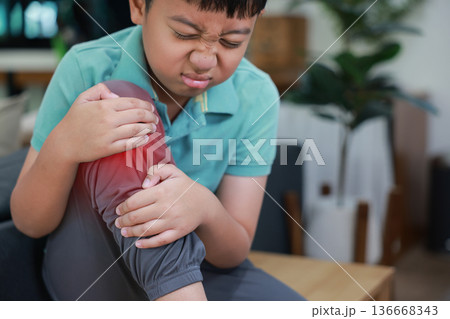 Asian boy showing painful face expression holding red inflamed knee from joint injury or accident. Pediatric healthcare concept of childhood bone problems, growth pain, and orthopedic treatment. 136668343