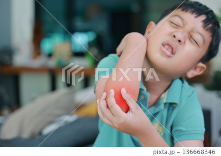 Asian boy suffering from elbow pain with red highlight on painful area, child having arm muscle injury, joint inflammation, accidental bone fracture or skin irritation problem at home. Asian boy suffering from elbow pain with red highlight on painful area, child having arm muscle injury, joint inflammation, accidental bone fracture or skin irritation problem at home. 136668346