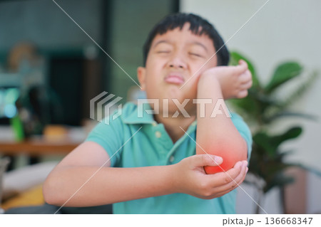 Asian boy suffering from elbow pain with red highlight on painful area, child having arm muscle injury, joint inflammation, accidental bone fracture or skin irritation problem at home. 136668347