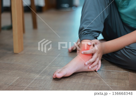 Close up of child's hands holding ankle with red highlight on painful area, Asian boy suffering from ankle sprain, foot injury, ligament strain or bone inflammation sitting on floor. 136668348