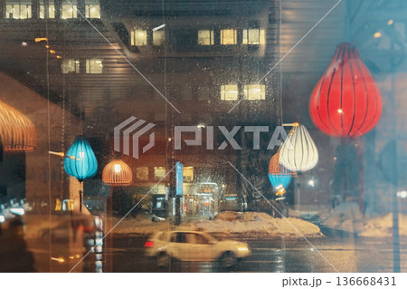 Colorful lanterns hanging inside cafe window with city street reflection and cars passing at night in urban winter scene. Night city lifestyle, window reflections, urban background Colorful lanterns hanging inside cafe window with city street reflection and cars passing at night in urban winter scene. Night city lifestyle, window reflections, urban background 136668431