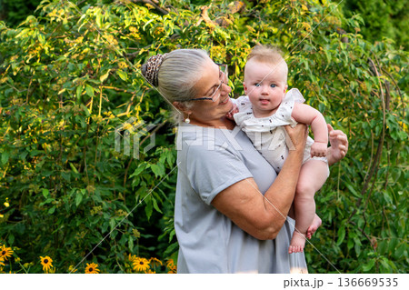 Grandmother holding smiling baby outdoors in lush garden Grandmother holding smiling baby outdoors in lush garden 136669535