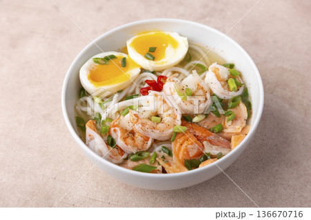 Asian dish with broth, noodles, tiger shrimp, fish, egg and green onions. Traditional Asian soup in a white deep plate. Selective focus, close-up. 136670716