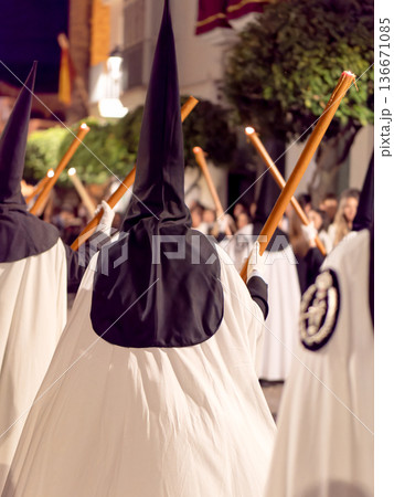 Nazarenes in white tunics and black hoods walking in a night procession with candles. 136671085
