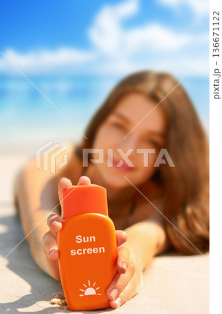 Portrait of gorgeous young woman smiling while relaxing on beach and holding sunscreen bottle 136671122