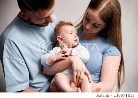 Family poses with baby in studio setting during daytime, showing warmth and connection in their interaction and expressions while holding the infant in their arms Family poses with baby in studio setting during daytime, showing warmth and connection in their interaction and expressions while holding the infant in their arms 136671818