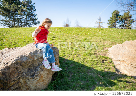 Little girl sitting on decorative stones blowing soap bubbles in city park. Spring childhood leisure and joyful outdoor play. 136672023