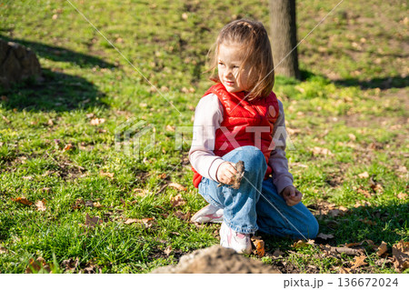 Little girl exploring nature in city park during spring walk. Curious child interacting with soil, leaves and landscape in outdoor environment. 136672024