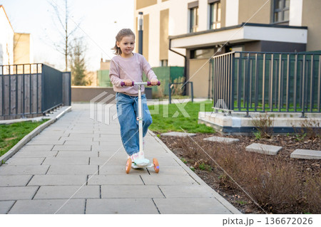 Little girl riding scooter in residential courtyard during spring. Active outdoor play and everyday childhood lifestyle in urban environment. 136672026