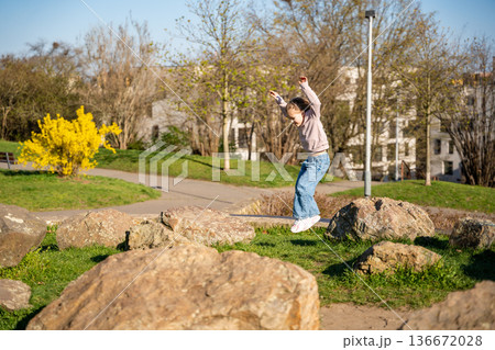 Little girl jumping on decorative stones in city park. Active spring play and outdoor childhood lifestyle. 136672028