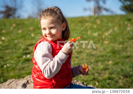 Little girl sitting on decorative stones blowing soap bubbles in city park. Spring childhood leisure and joyful outdoor play. 136672038