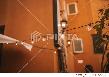 An outdoor patio at night illuminated by warm string lights and hanging lanterns. Woven furniture including a couch and chairs are arranged around tables on a tiled floor. Potted plants and trees An outdoor patio at night illuminated by warm string lights and hanging lanterns. Woven furniture including a couch and chairs are arranged around tables on a tiled floor. Potted plants and trees 136672257