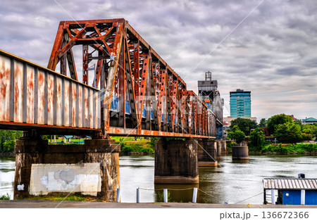 Clinton Presidential Park Bridge in Little Rock, Arkansas. Historic rusty steel truss railway bridge spans the Arkansas River with the River Market Tower in the background under a cloudy sky 136672366