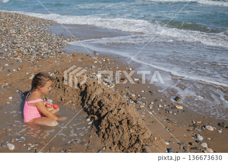 Happy Little Girl in Pink Swimsuit Building Sand Castle Moat on a Pebble Beach with Waves and Sea Foam in Summer Sunlight 136673630