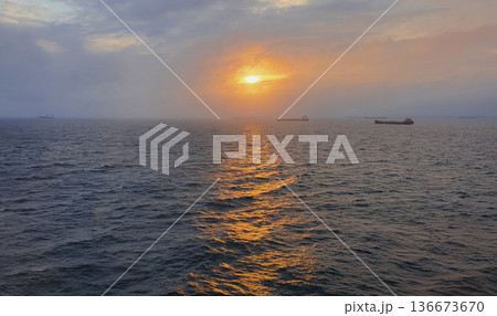 Dramatic pre-sunset seascape golden sunlight breaking through dense gray clouds, illuminating rippling dark blue water distant cargo ships on horizon 136673670