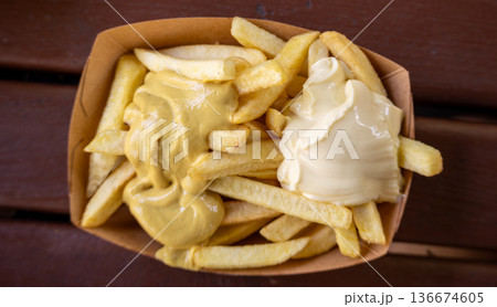 Berlin, germany, august 14, 2023. Golden french fries in a paper tray topped with creamy mayonnaise and yellow mustard, offering a popular fast food snack 136674605