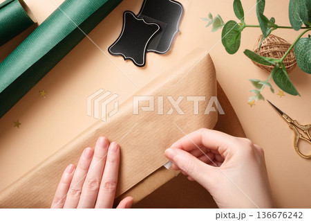 Top view of hands wrapping a gift in brown kraft paper. Festive background with green paper rolls, twine, gold scissors. Top view of hands wrapping a gift in brown kraft paper. Festive background with green paper rolls, twine, gold scissors. 136676242
