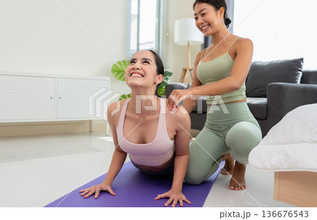 Two smiling sporty girls practice yoga with partner at home. Trainer helps student to do Bhujangasana exercise or Cobra pose Two smiling sporty girls practice yoga with partner at home. Trainer helps student to do Bhujangasana exercise or Cobra pose 136676543