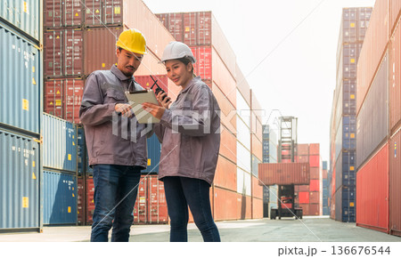 Two logistics workers in hard hats and uniforms using digital tablet and walkie talkie for loading containers control at cargo shipping container yard Two logistics workers in hard hats and uniforms using digital tablet and walkie talkie for loading containers control at cargo shipping container yard 136676544