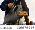 Barista in gray apron carefully pouring hot water from black kettle into glass pour over coffee dripper which sit top wooden stand for manual coffee brewing. Brewed coffee drip into clear glass below 136676546