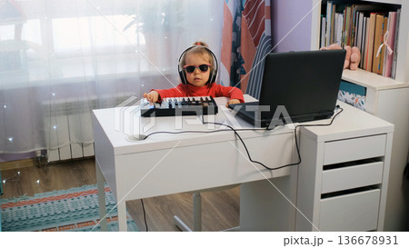 Cute little girl enjoys making music on a midi keyboard at home. She wears headphones and sunglasses while moving her head to the rhythm Cute little girl enjoys making music on a midi keyboard at home. She wears headphones and sunglasses while moving her head to the rhythm 136678931