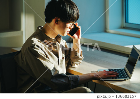 パソコンを見ながらスマホで話す若い男性 パソコンを見ながらスマホで話す若い男性 136682157