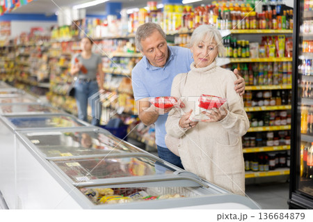 Elderly couple choosing foods in glass display case in supermarket Elderly couple choosing foods in glass display case in supermarket 136684879