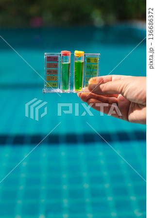 Pool water testing kit measuring pH and chlorine levels during routine swimming pool maintenance with clear blue water in background 136686372