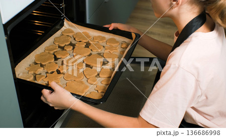 A teenage girl wearing an apron places a tray of raw ginger cookies into the oven, baking homemade bakery A teenage girl wearing an apron places a tray of raw ginger cookies into the oven, baking homemade bakery 136686998